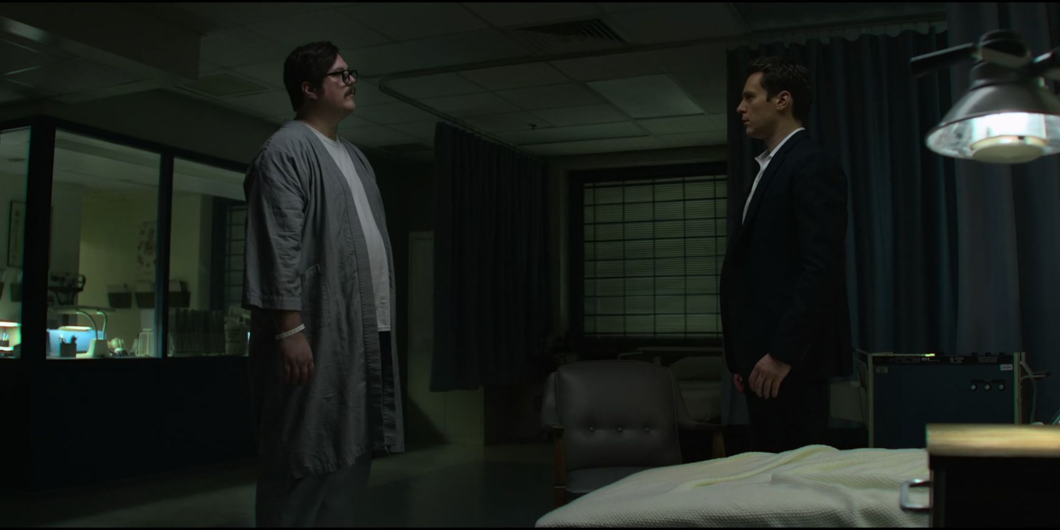 Cameron Britton as Ed Kemper stands in a robe and stares at Jonathan Groff as Holden Ford in a suit in Mindhunters