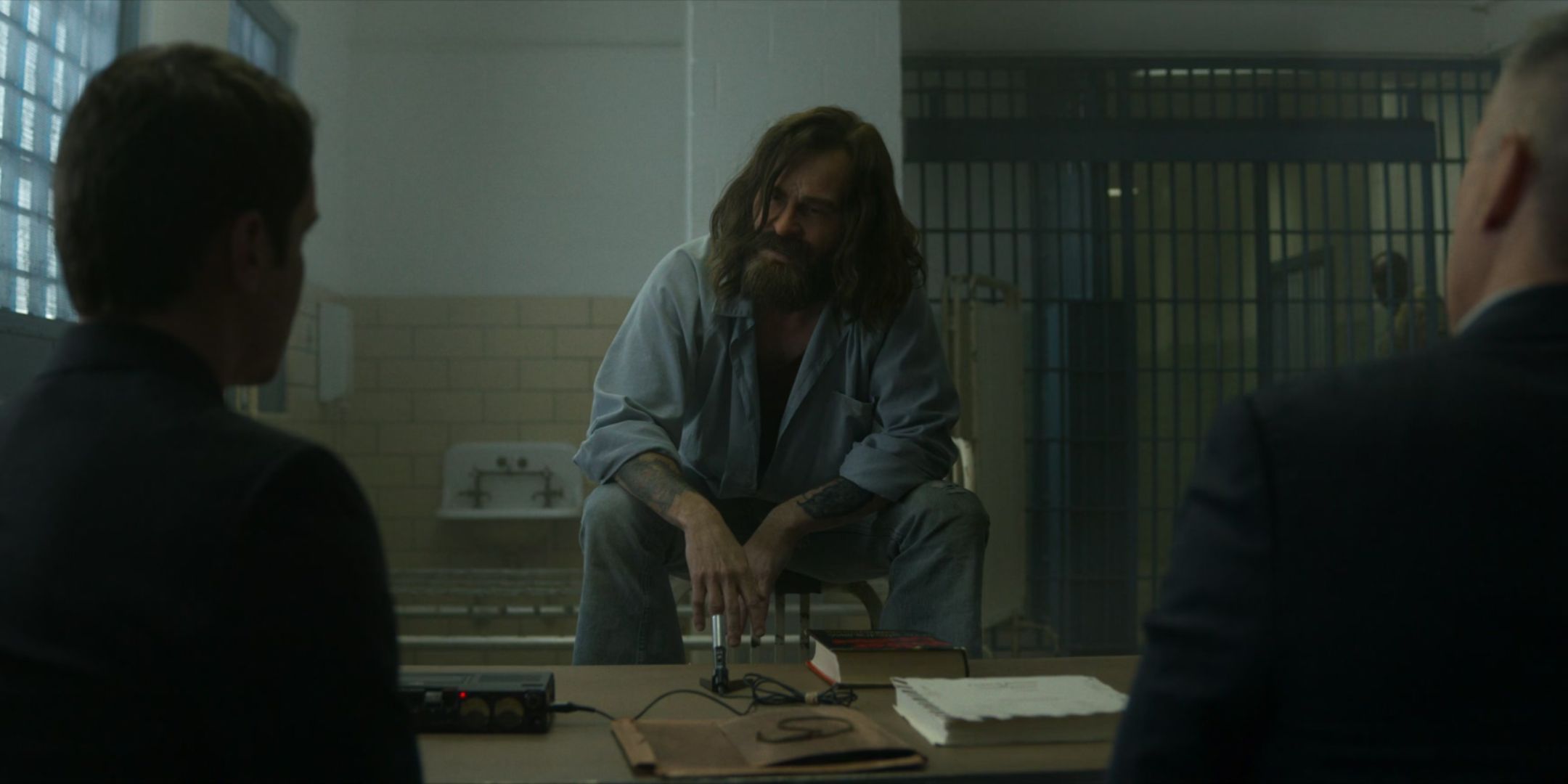 Damon Herriman as Charles Manson sits on the back of a chair in prison and talks to Jonathan Groff as Holden Tench and Holt McCallany as Bill Tench in Mindhunter