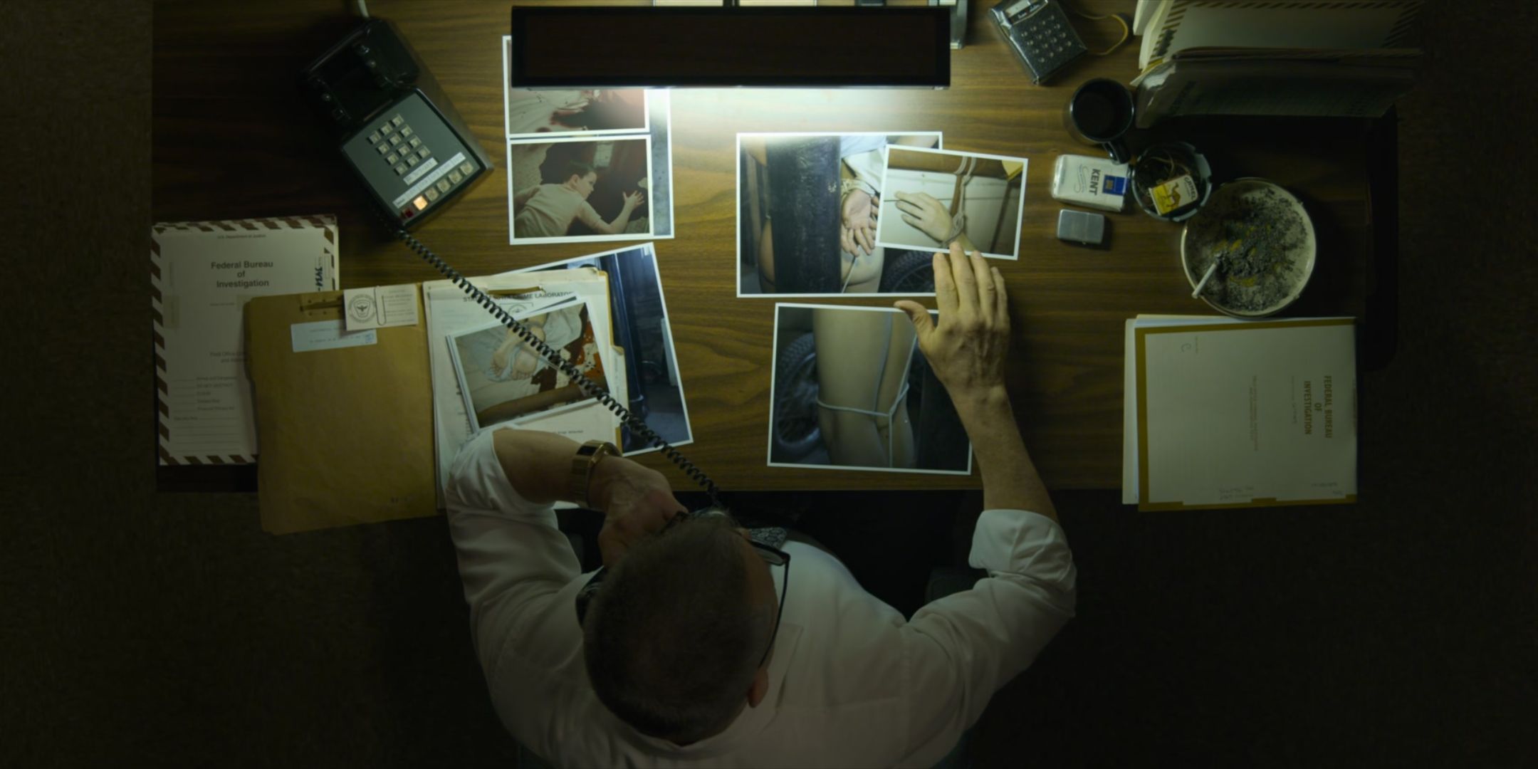 Holt McCallany as Bill Tench sits at a desk and looks at photos of a murder in Mindhunter