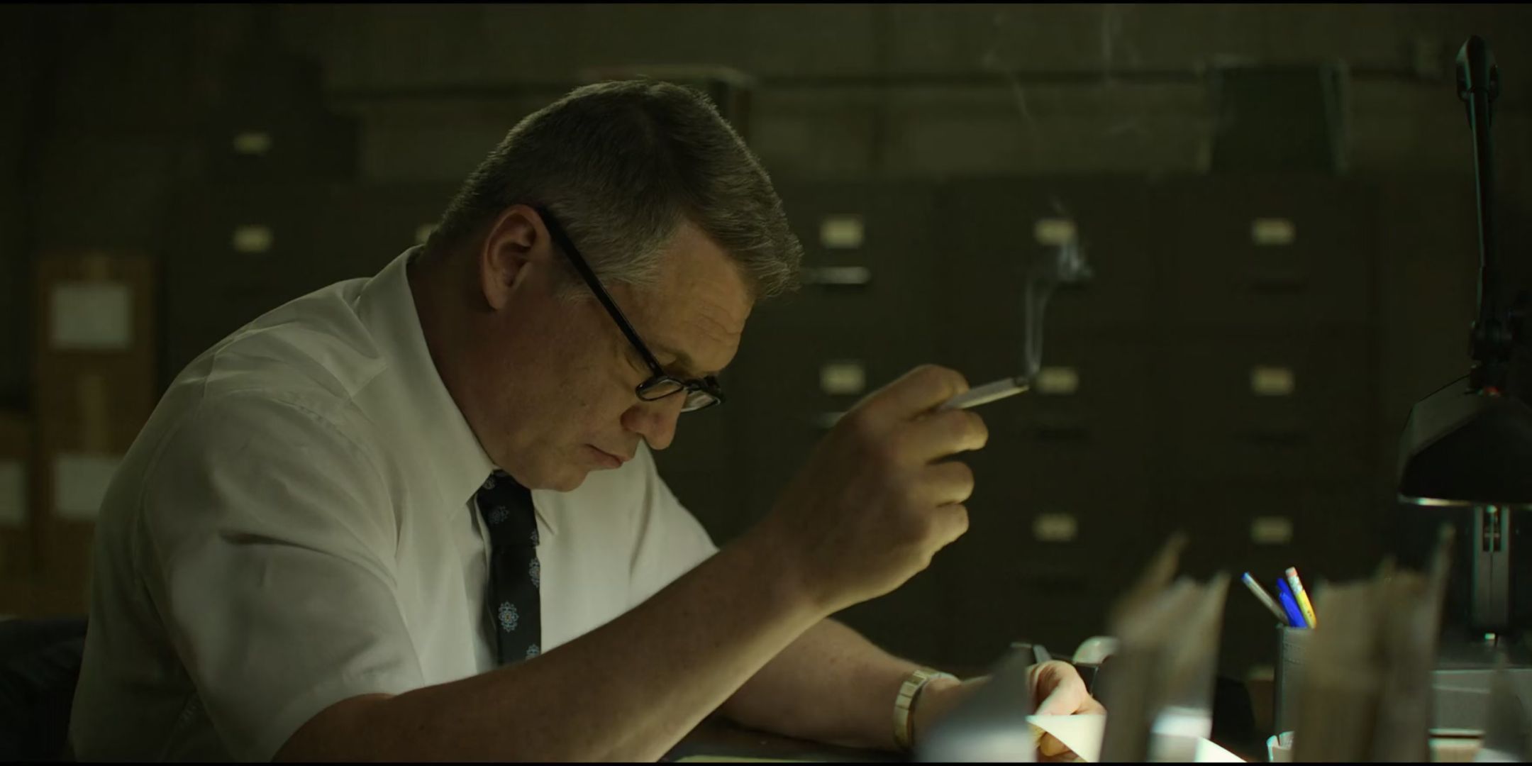 Holt McCallany as Bill Tench studies something in a shirt and tie while holding a cigarette in Mindhunter