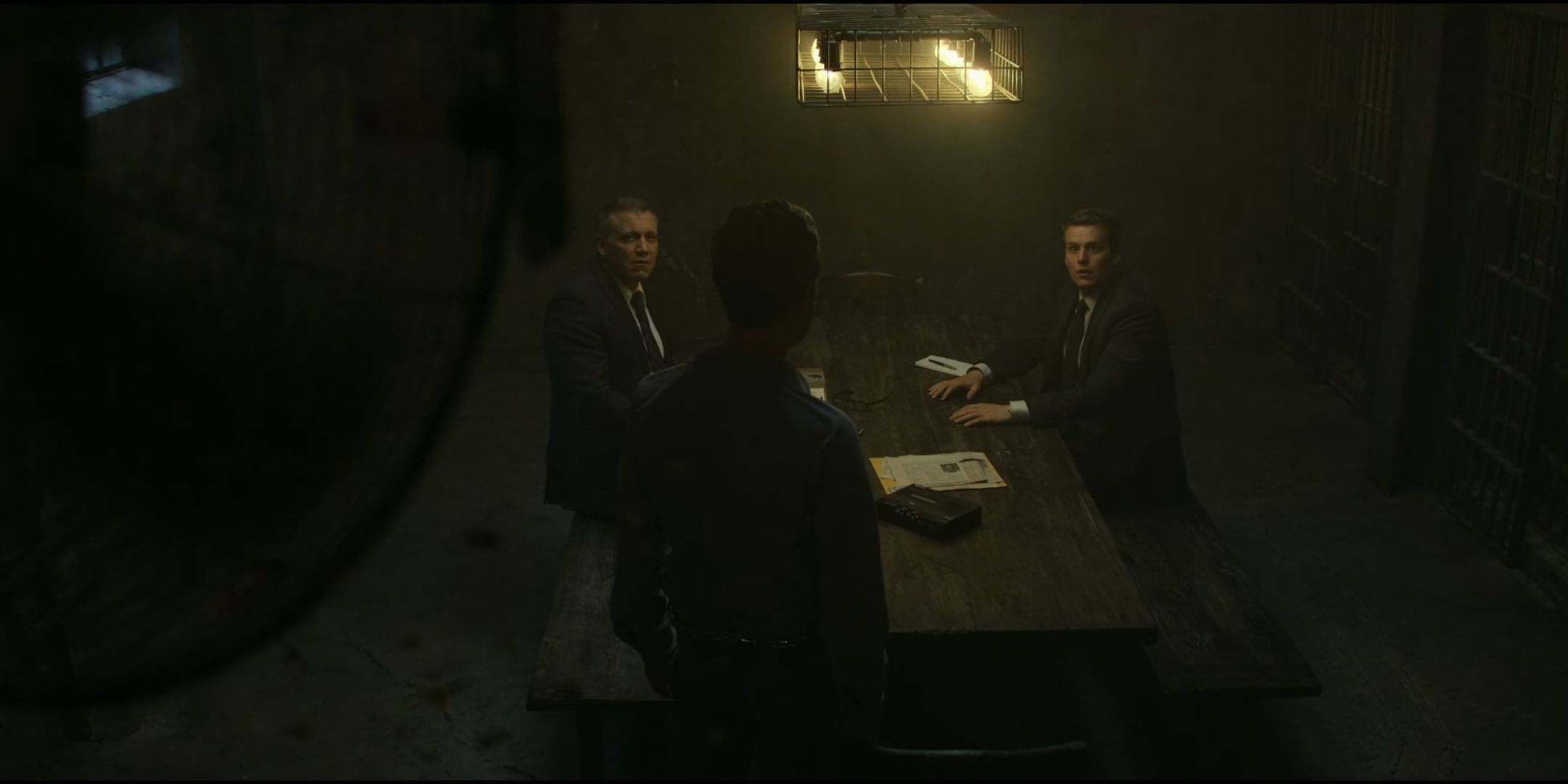 Jack Erdie as Richard Speck looks at Jonathan Groff as Holden Ford and Holt McCallany as Bill Tench at a desk with a fan in the background in Mindhunter