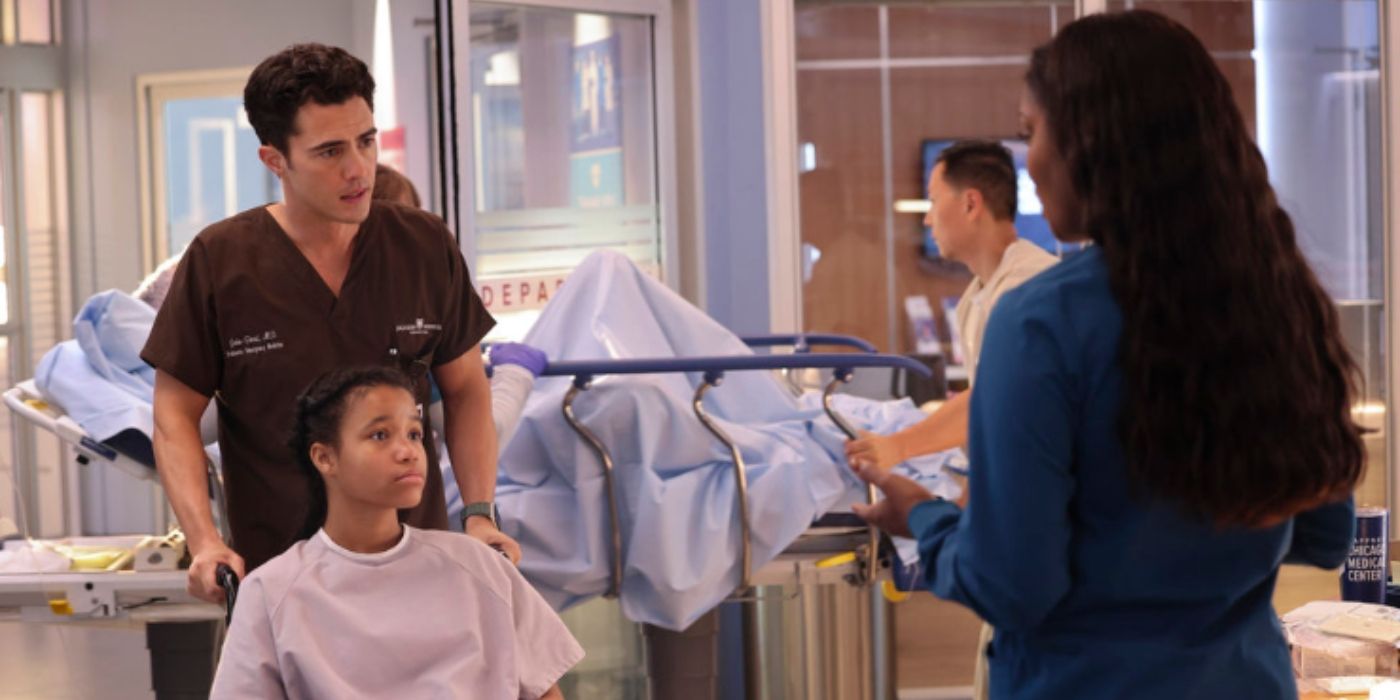 Darren Barnet as John Frost pushes a patient in a wheelchair while speaking to Marlyne Barrett as Maggie Lockwood who is facing away from the camera on Chicago Med