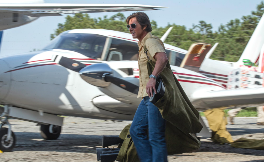 Barry Seal carrega sacos de armas em um aeroporto com um avião bimotor atrás dele em American Made.