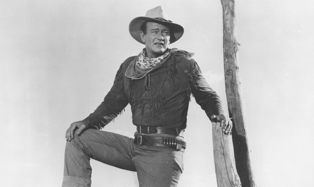 John Wayne como Hondo fica encostado em uma cerca com o chapéu voltado para o céu.