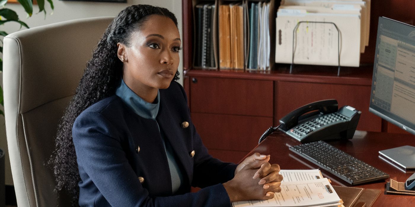 Andrea, played by Yaya DaCosta, sits at a desk in a blue blazer in the third season of The Lincoln Lawyer