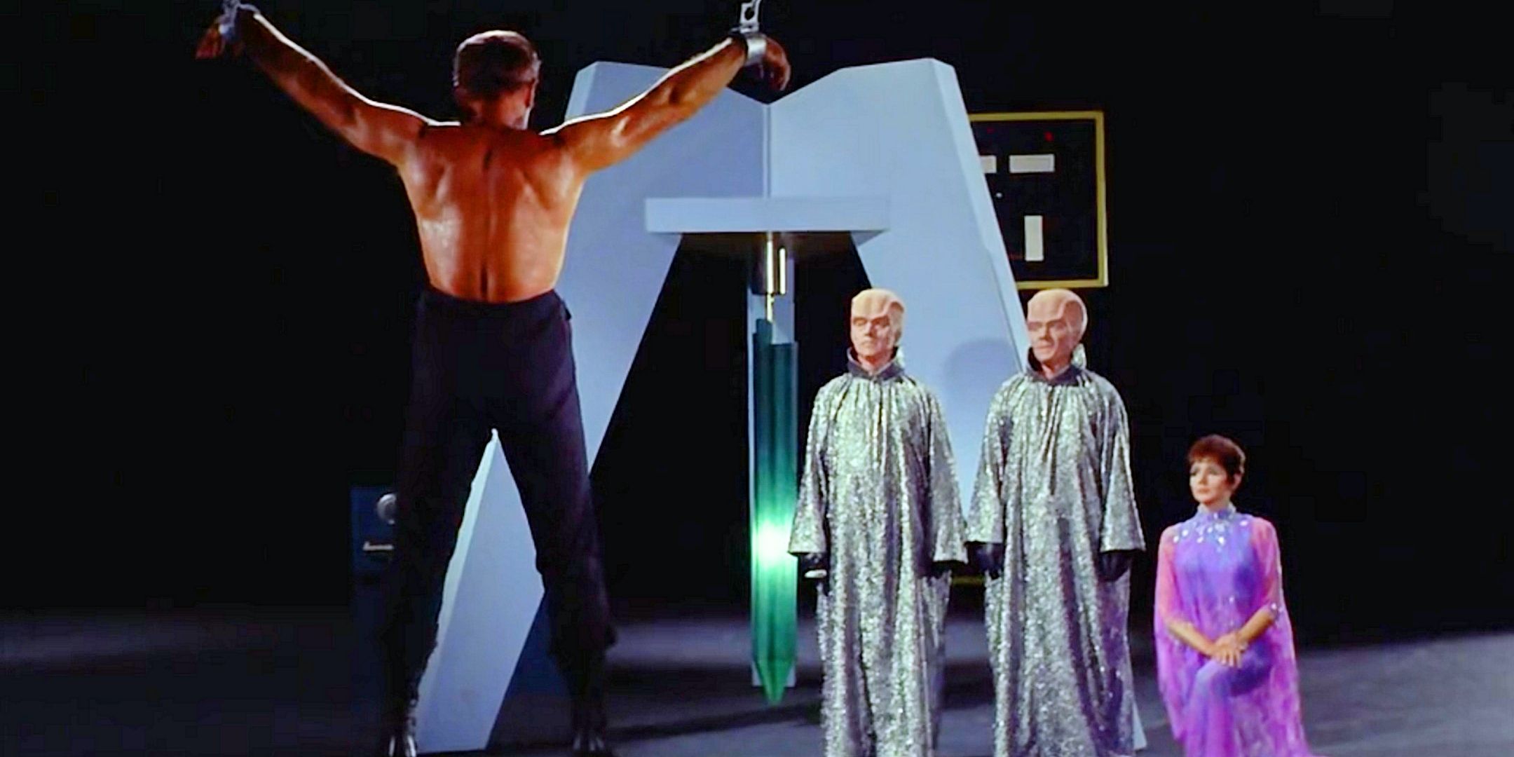 A shirtless Kirk, back to camera, is suspended by his limbs as two bal aliens in silver dresses torture him Star Trek The Original Series