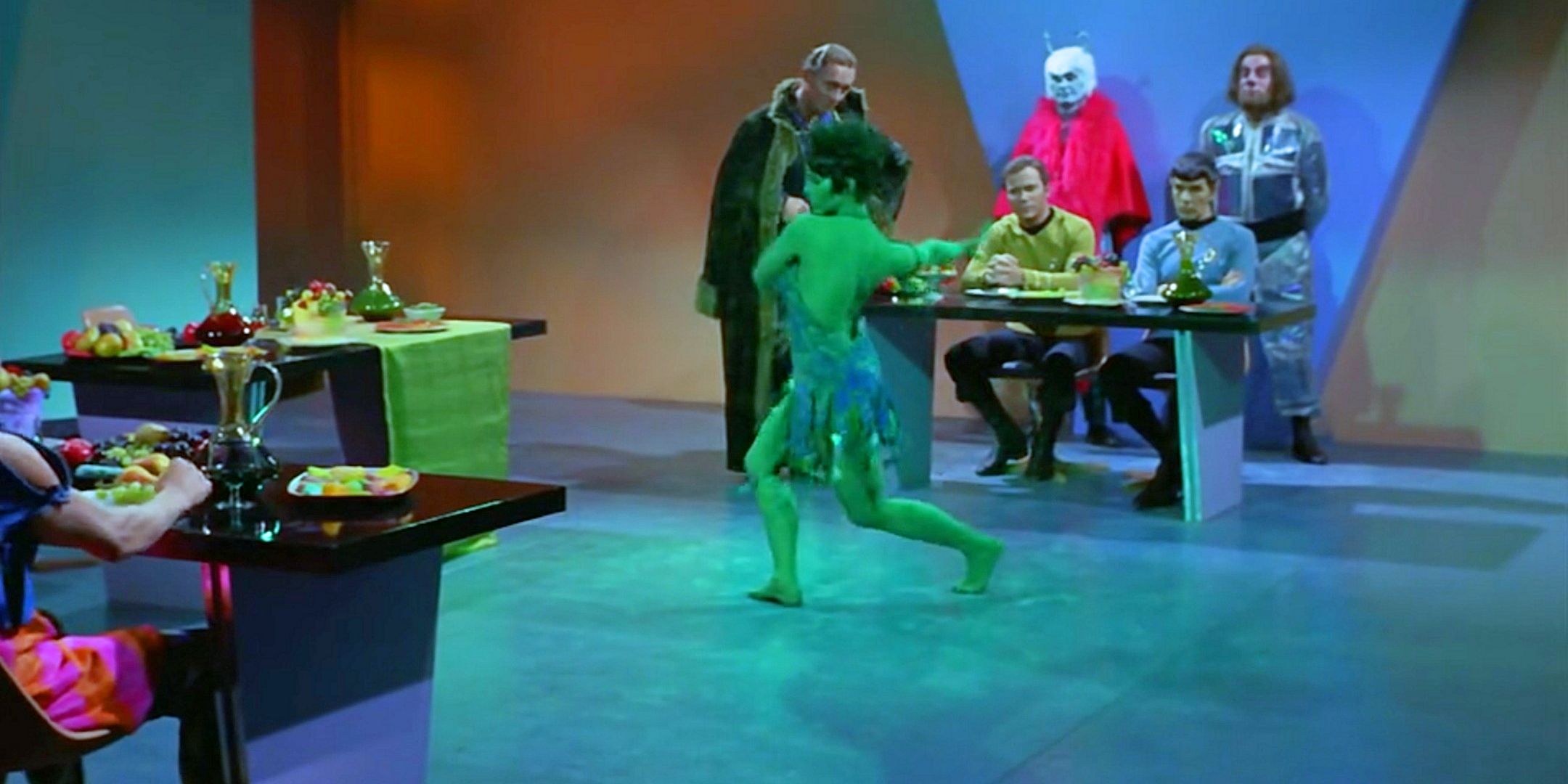 An Orian woman in a skimpy dress dances as ostentatious aliens stand by a table where Kirk and Spock sit in uniform and watch from Star Trek The Original Series