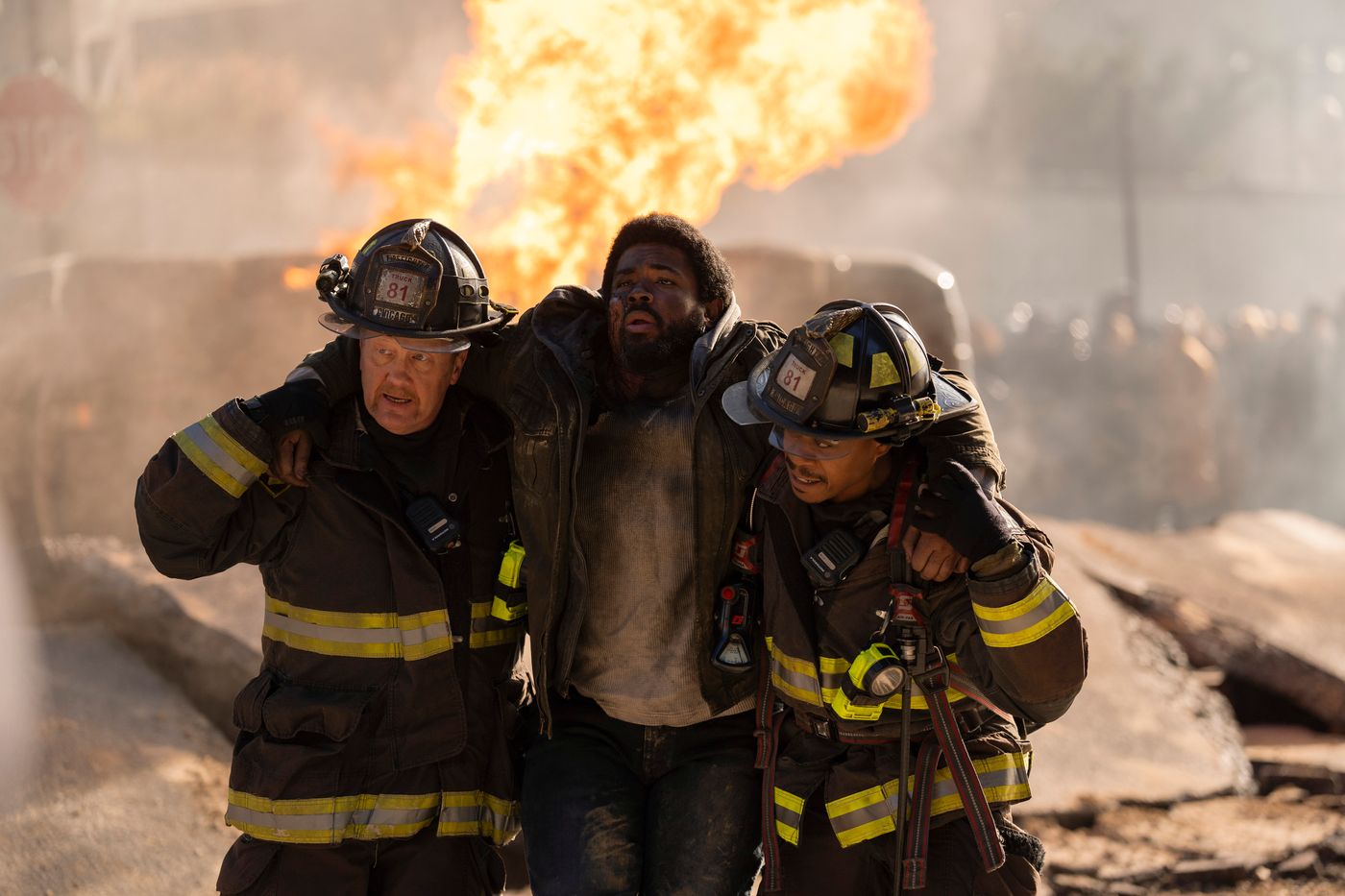 Mouch and Ritter, wearing firefighter gear, hold up an injured man between them on Chicago Fire