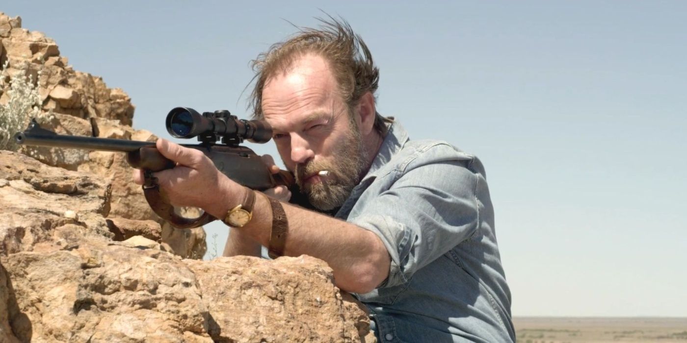 Hugo Weaving como Johnno aponta um rifle com um cigarro na boca em Mystery Road