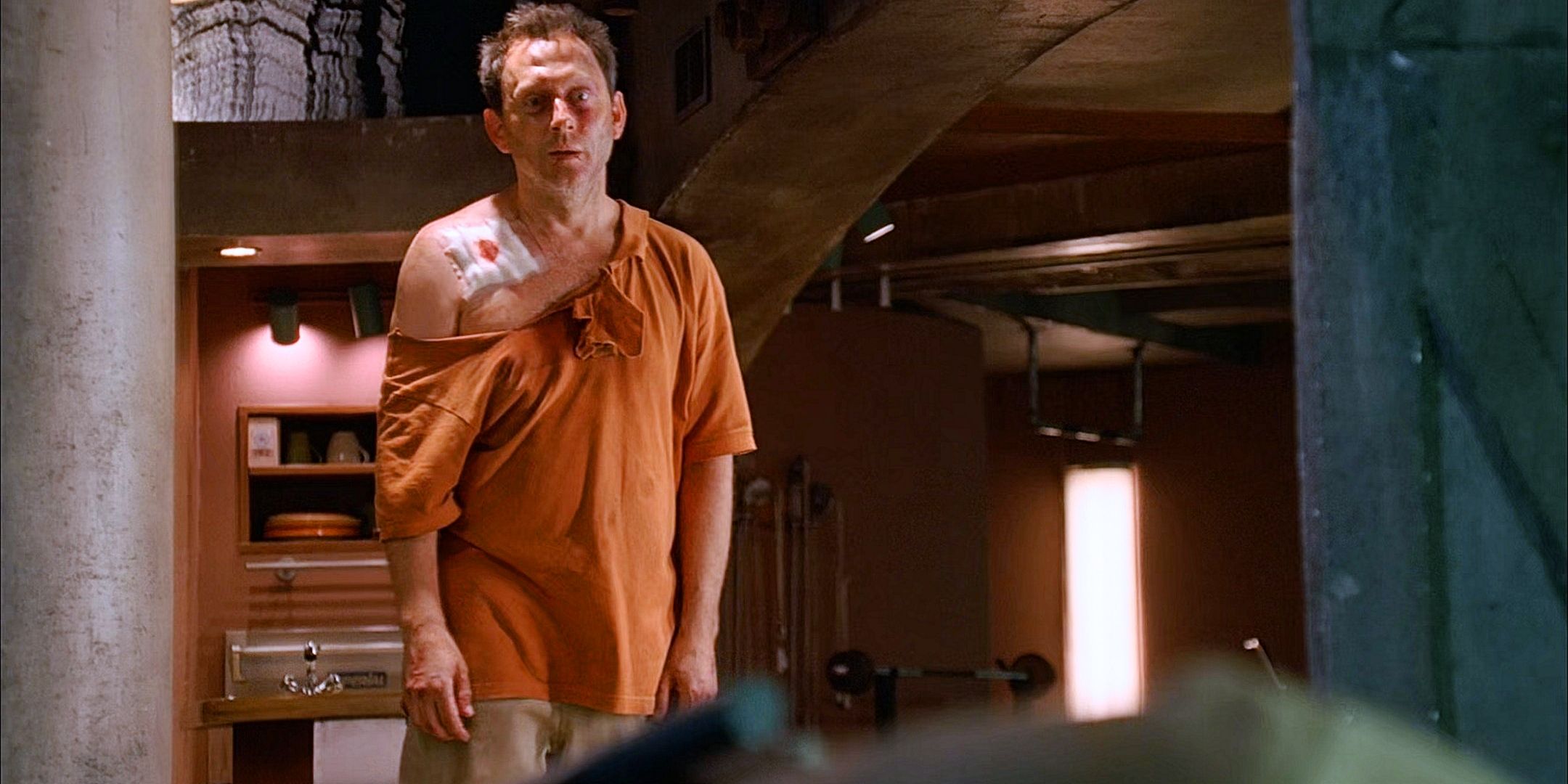 Ben Linus as Henry gale in a torn orange polo with bandaged shoulder and khakis standing in the hatch living area on the Lost series