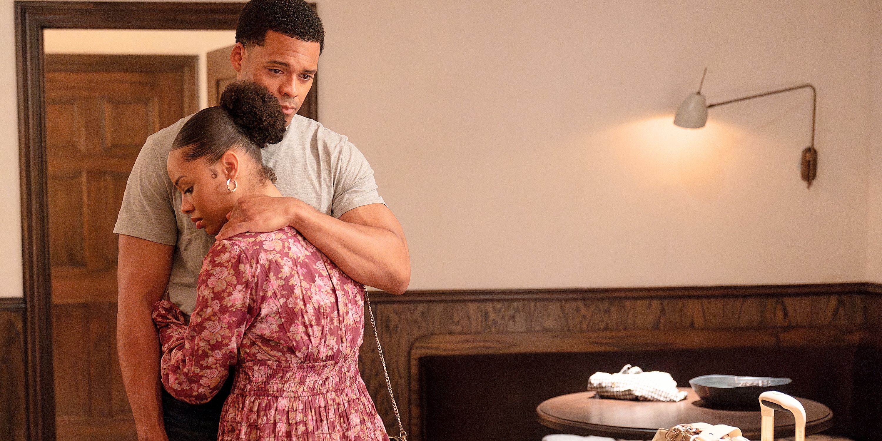 Miles Penn con ropa Civlian Hug Camilla, con un vestido de flores al lado del equipaje, con un brazo en una habitación de hotel del novato