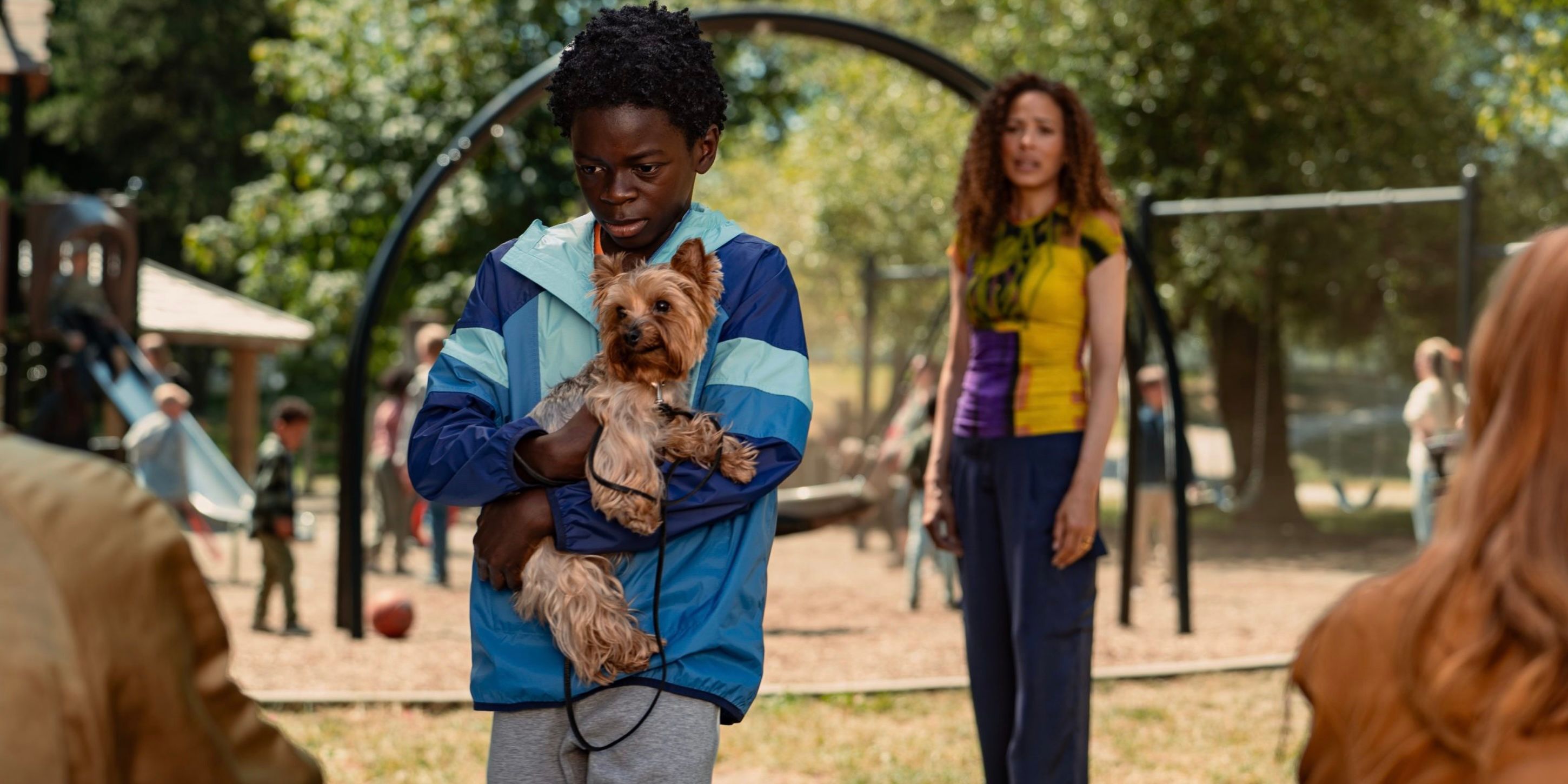 Sam holding a dog with Tai in the background at a playground on Yellowjackets
