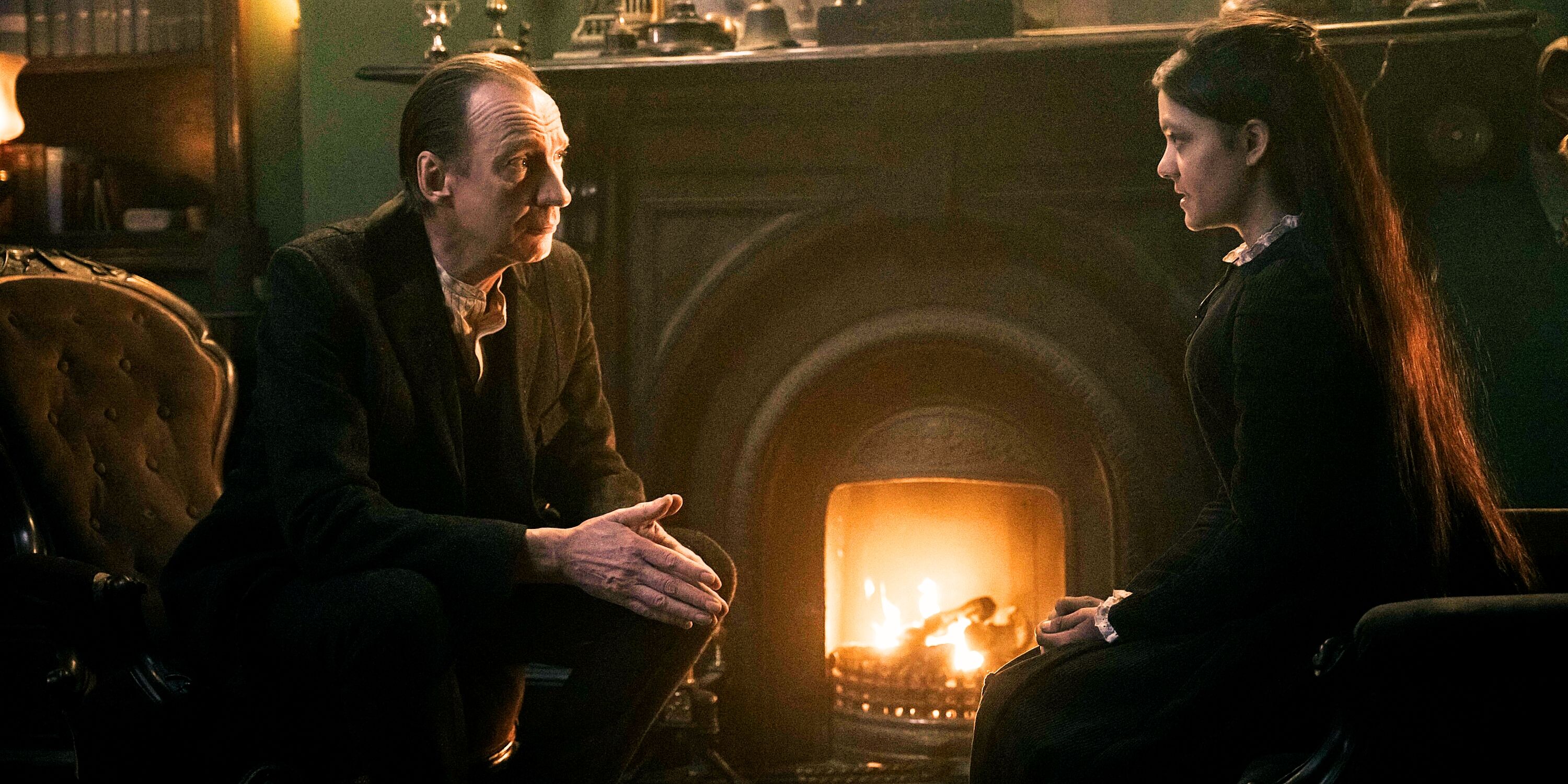 Sherlock Holmes in a brown coat faces Amelia Rojas in a black dress, both seated in front of a fireplace