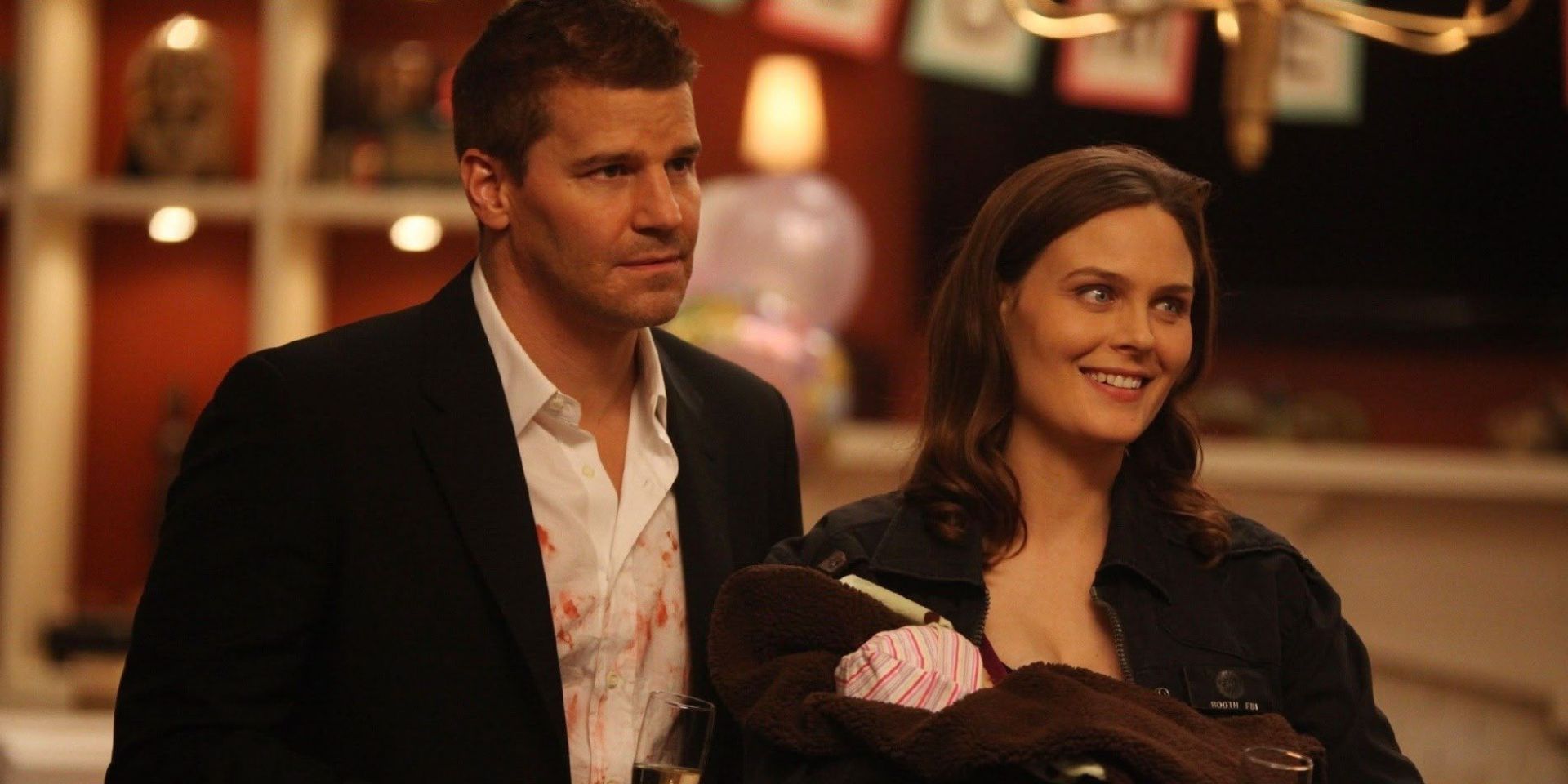 Booth (David Boreanaz) and Brennan stand together smiling, holding their baby in the TV show Bones