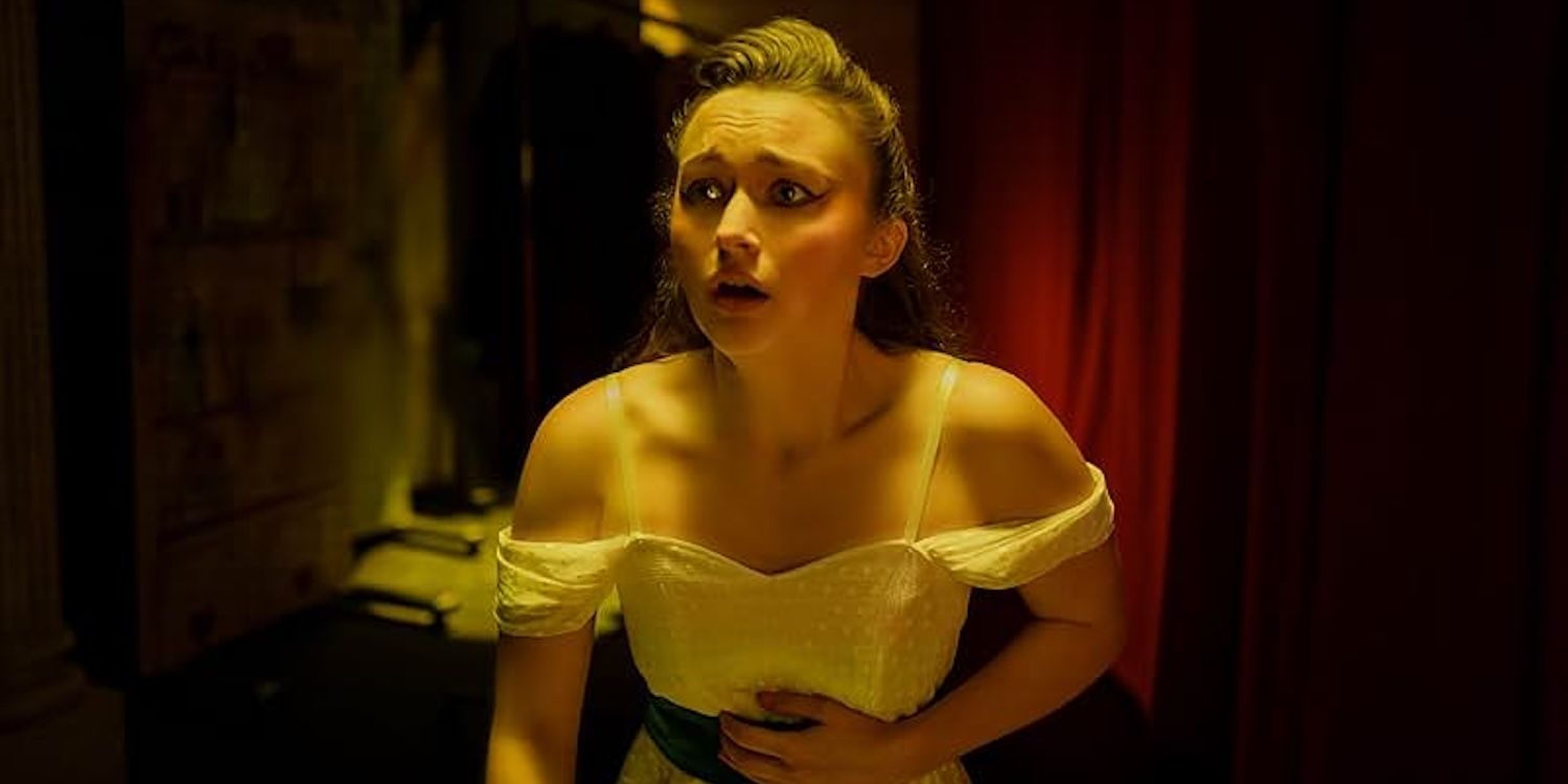 Lori Granger (India Fowler), wearing a white dress, clutches her stomach in horror behind a stage.