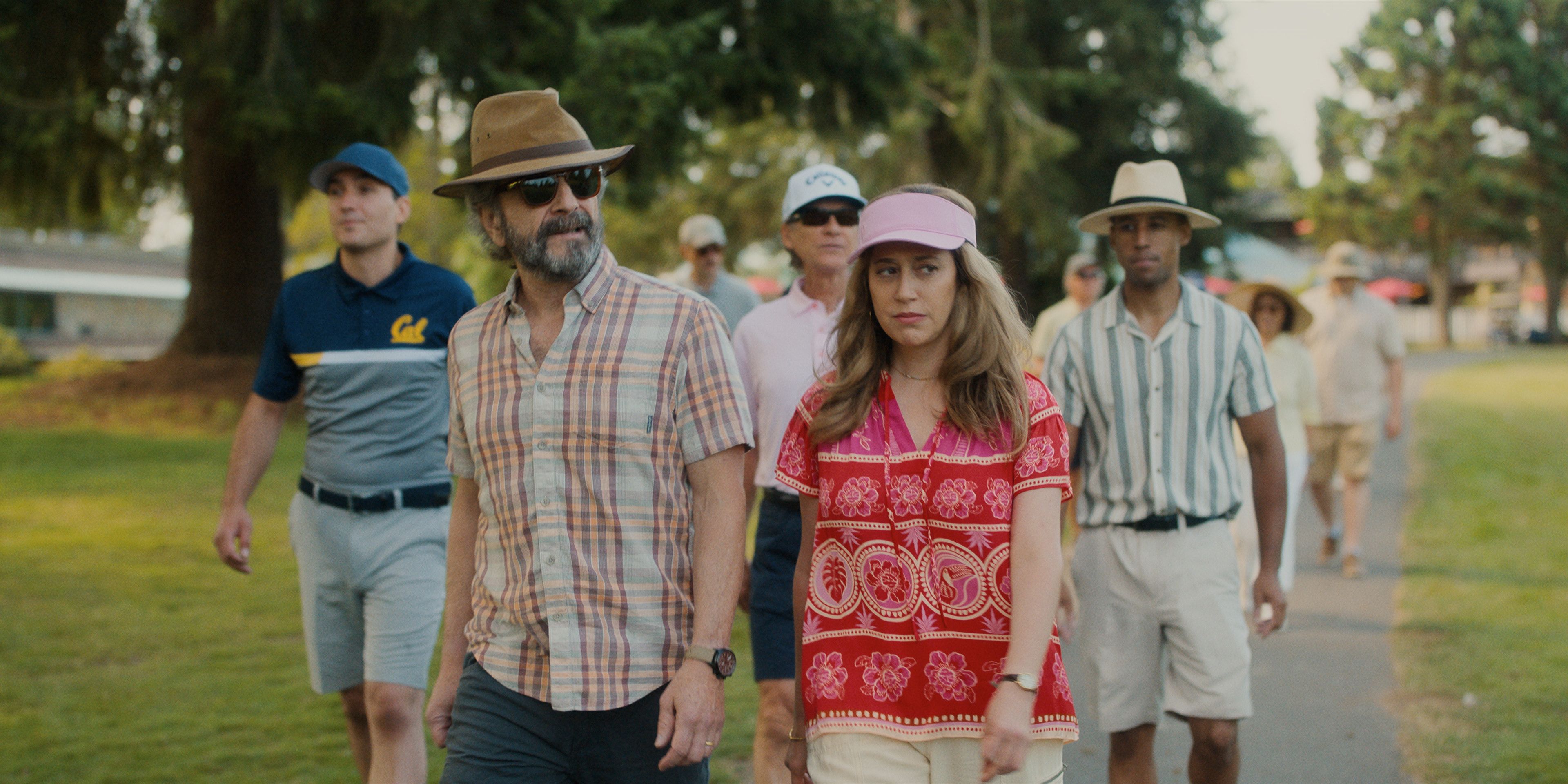 Mitts, in a jacket and a brown hat, walks with Elena on the golf course in the Apple Stick TV show