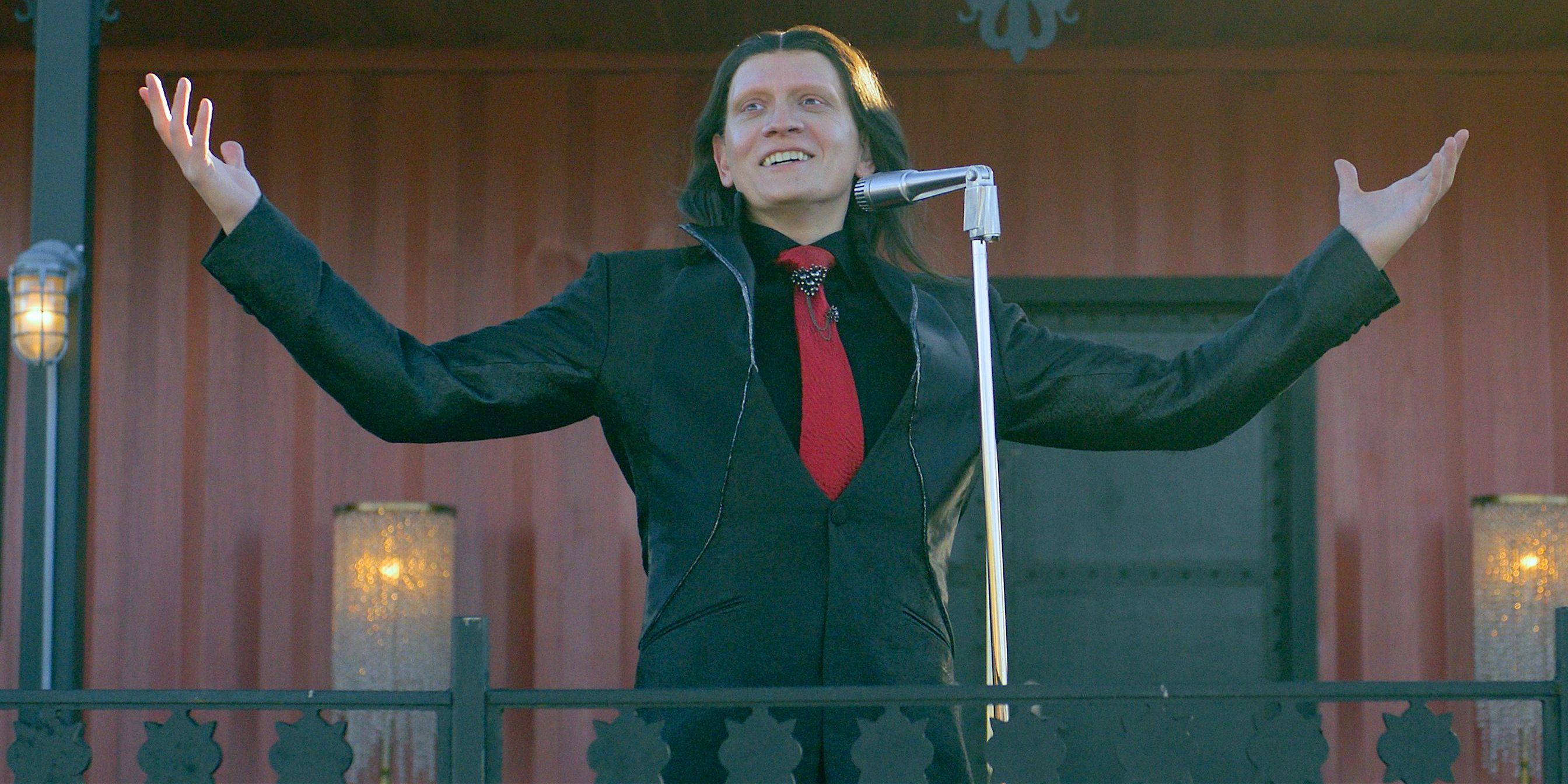 Calypso in a dark suit and red tie in front of a microphone from Twisted Metal