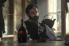 George Russell (Morgan Spector) sitting in at a table with a bottle in a saloon in Morenci in The Gilded Age Season 3.